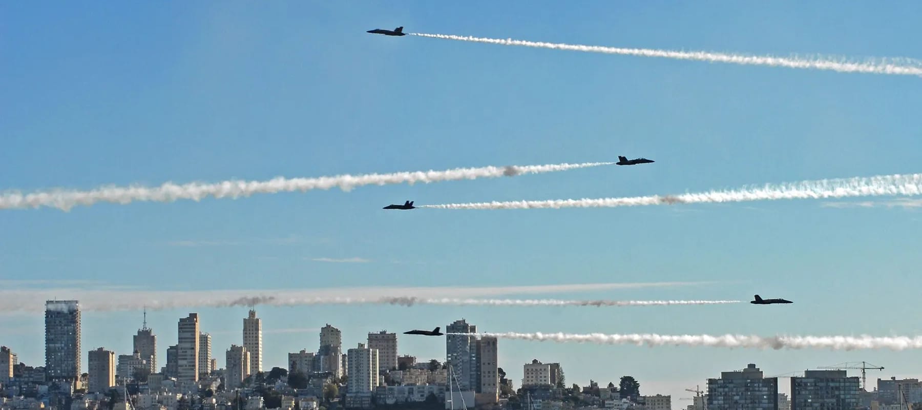 Six Blue Angels jets in formation trailing smoke above the San Francisco skyline, with boats on the water below
