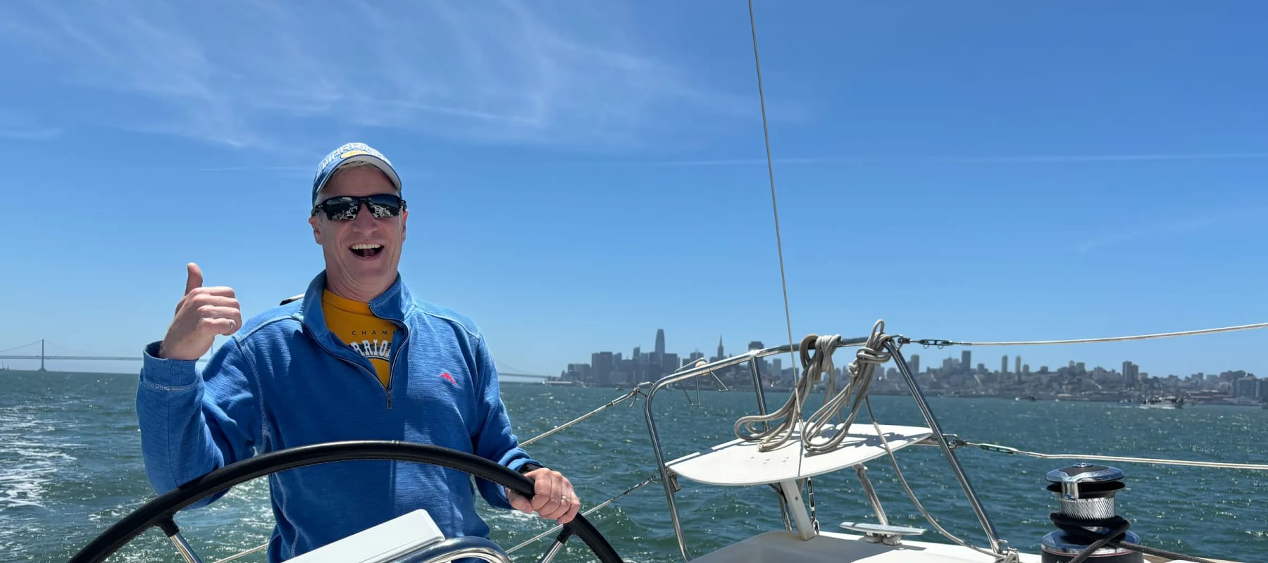 A guest at the wheel of a large sailboat on San Francisco Bay, grinning and giving a thumbs up, with the city skyline behind