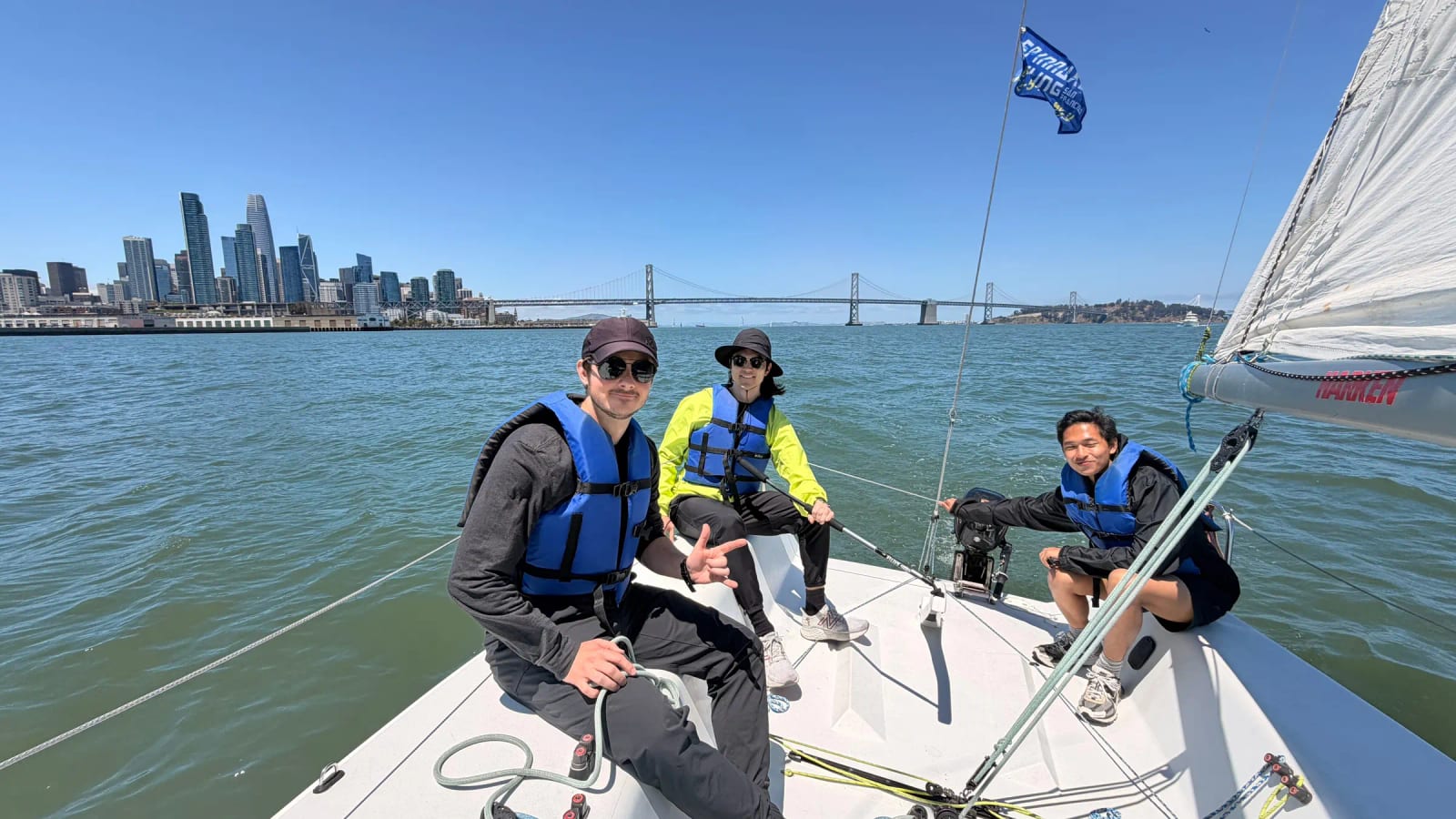 Three people sitting in the cockpit of a small keelboat on San Francisco Bay, one steering with a tiller, with the city skyline and Bay Bridge behind them