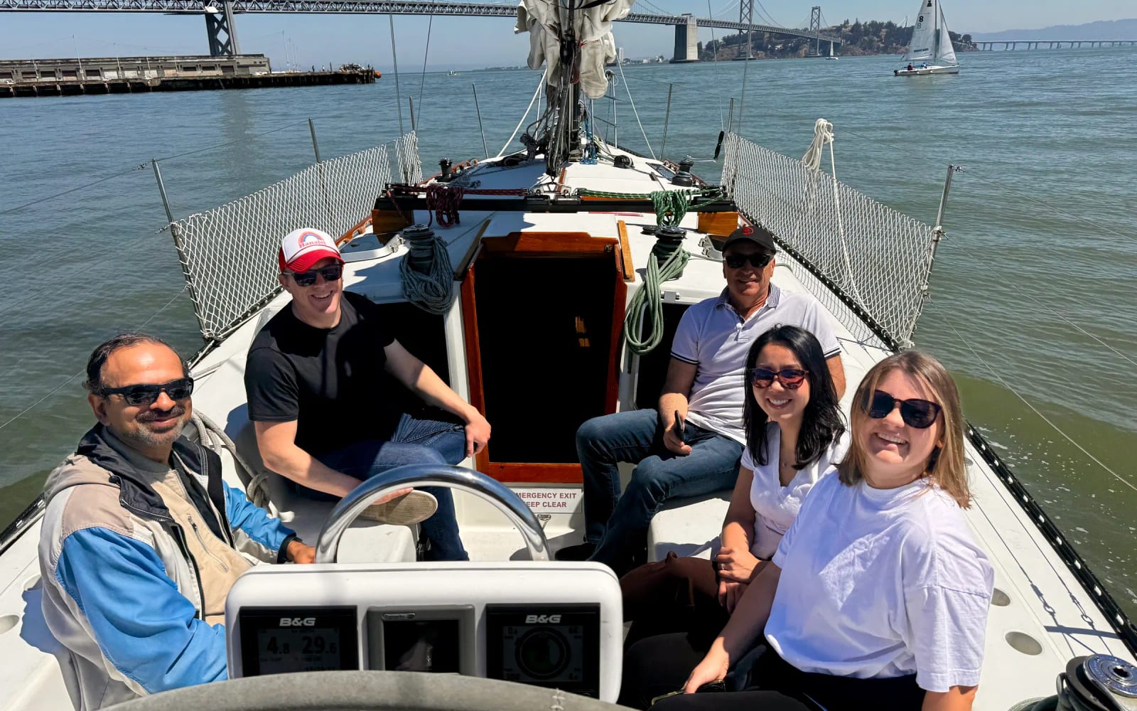 Guests seated around the wheel in the large cockpit of a fifty foot sailboat, with the Bay Bridge behind