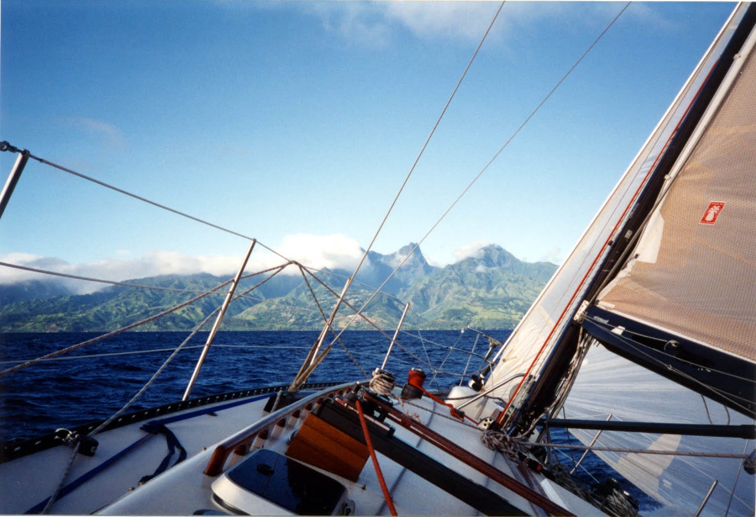 The deck of a racing sailboat heeled under full sail on deep blue ocean, with steep green volcanic peaks on the horizon
