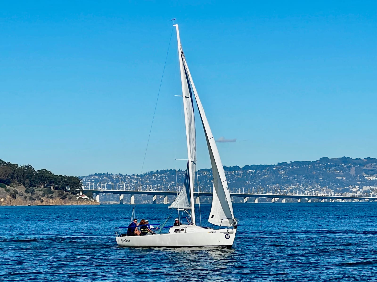 A small white sailboat sailing on deep blue water under a cloudless sky, with Yerba Buena Island, the Bay Bridge and the East Bay hills behind