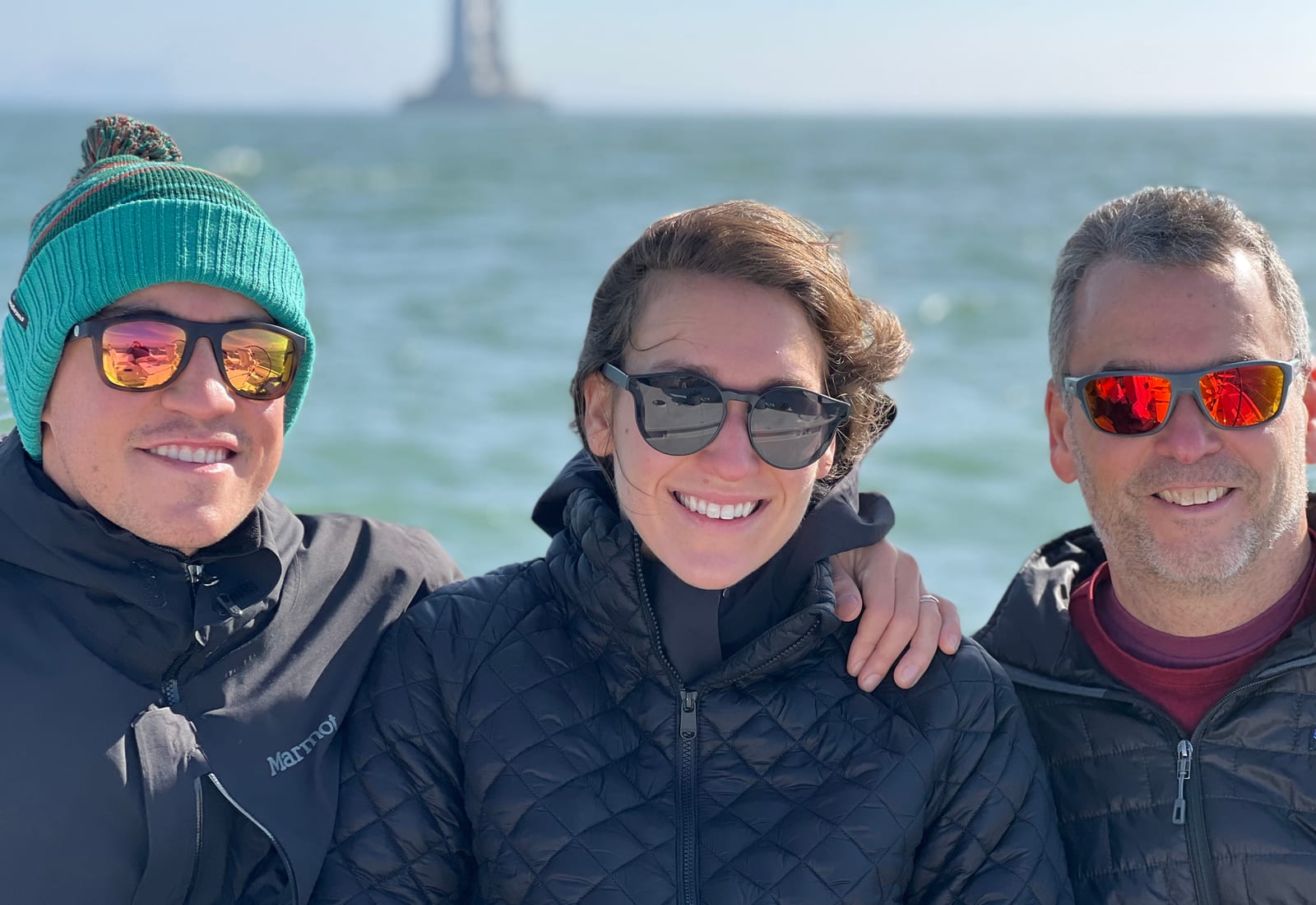 Three friends in a knitted beanie, sunglasses and padded jackets smiling on a sailboat, with a tower of the Bay Bridge in the haze behind