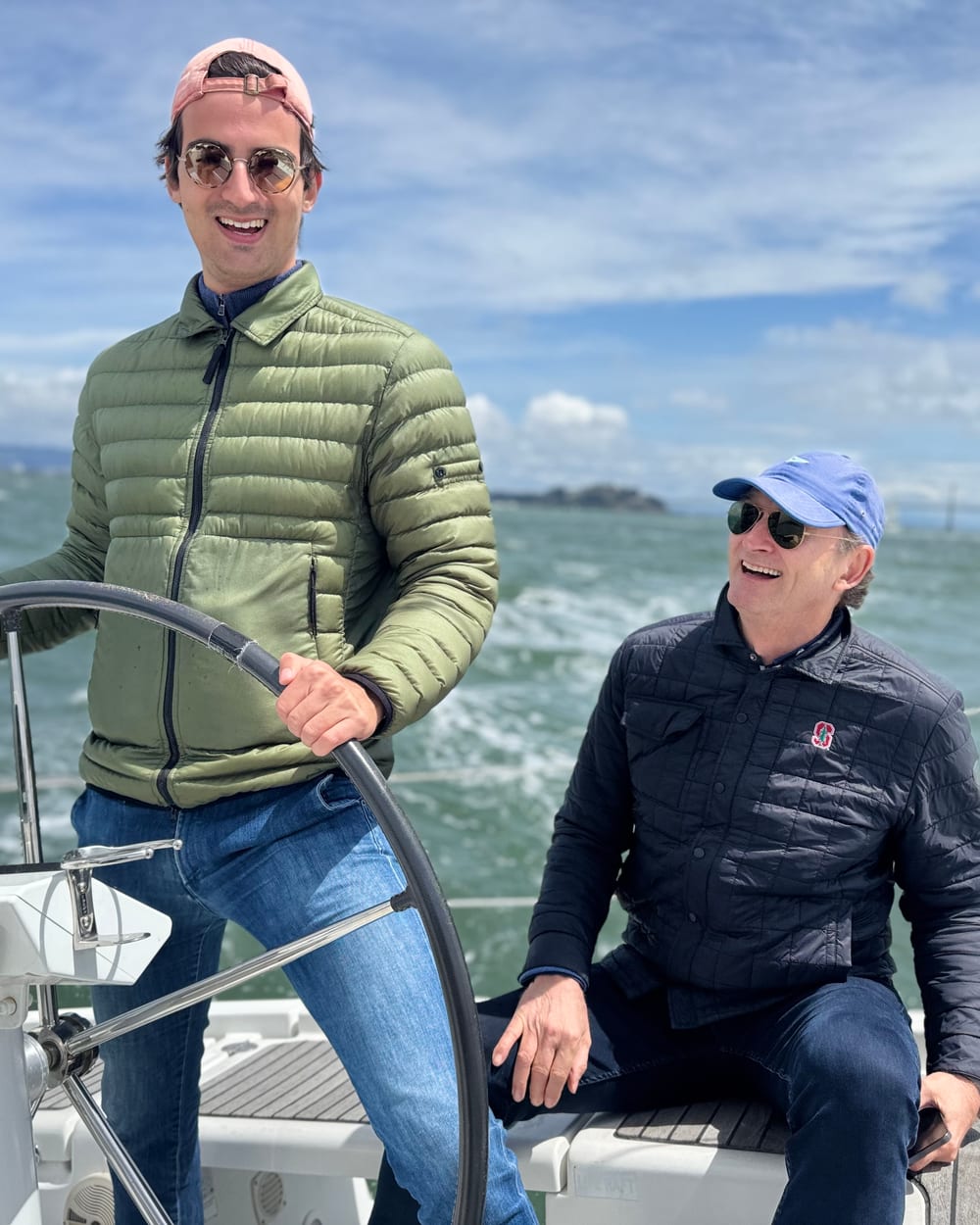 A guest in a green puffer jacket steering a sailboat with a big wheel on a sunny day, with another guest in a cap and quilted jacket beside him
