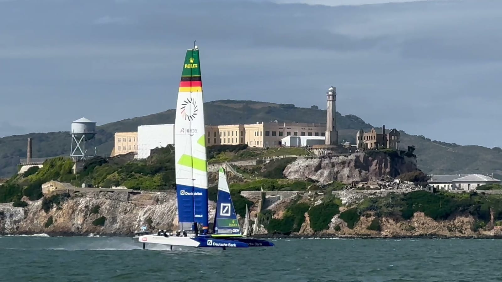 A SailGP F50 catamaran with a white, green and blue wing sail racing past Alcatraz Island