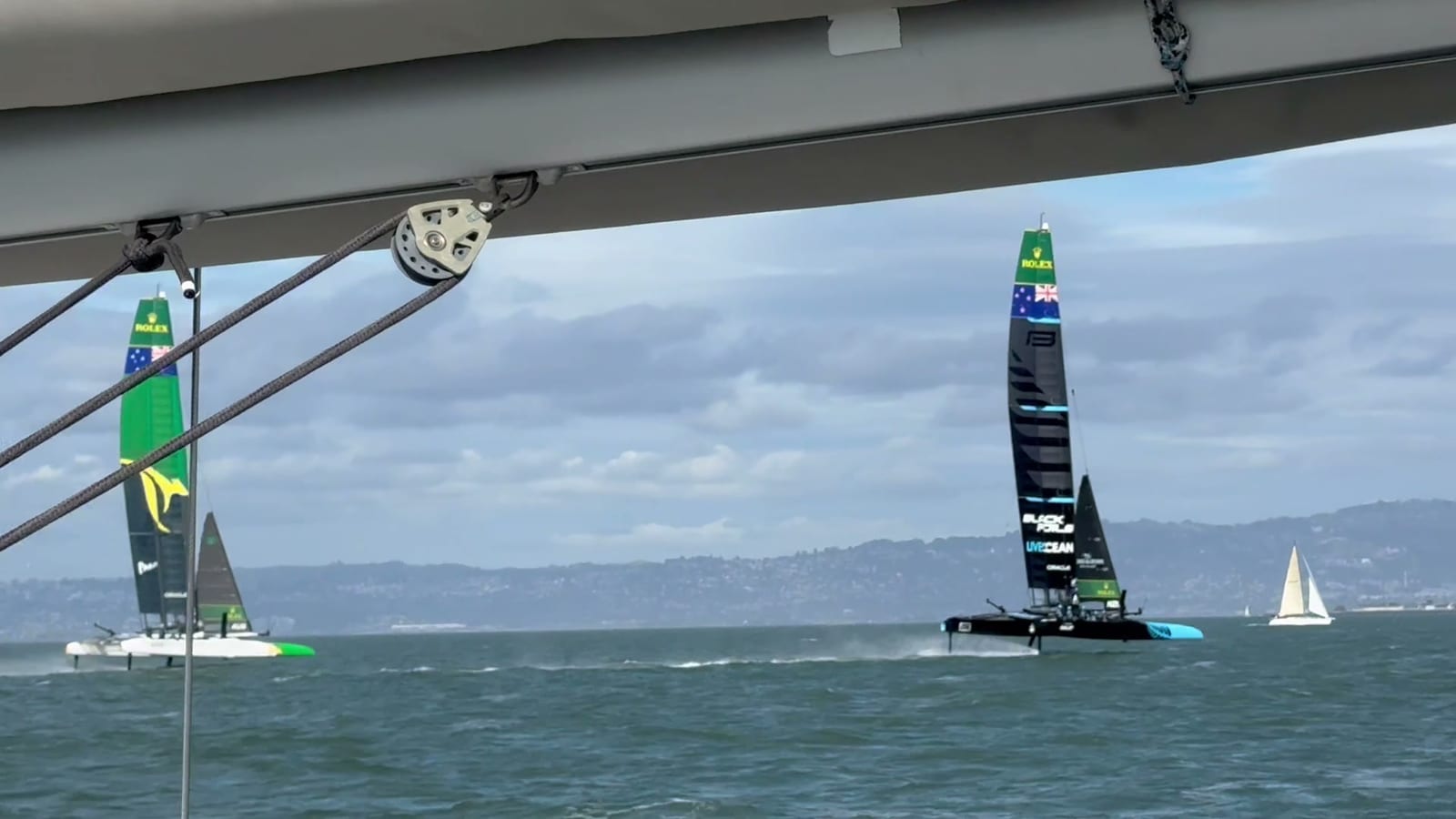 Two SailGP F50 catamarans foiling across the Bay, seen from under the boom and rigging of a sailboat