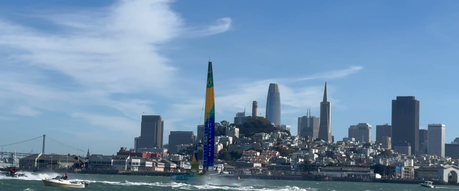 A SailGP F50 with a green and yellow wing sail racing in front of Coit Tower and the San Francisco skyline