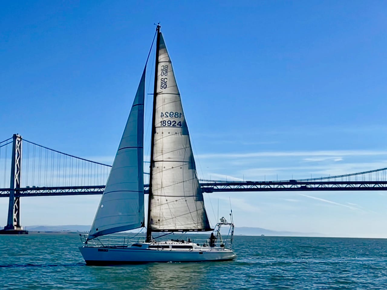 Yukon Jack, a white sailboat with the sail number 18924 on her mainsail, sailing on calm blue water in front of the Bay Bridge
