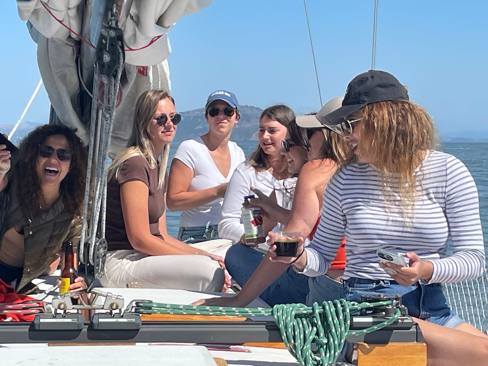 A group of women sitting on the deck of a sailboat beside the mast, laughing and holding drinks, with the Marin hills across the water
