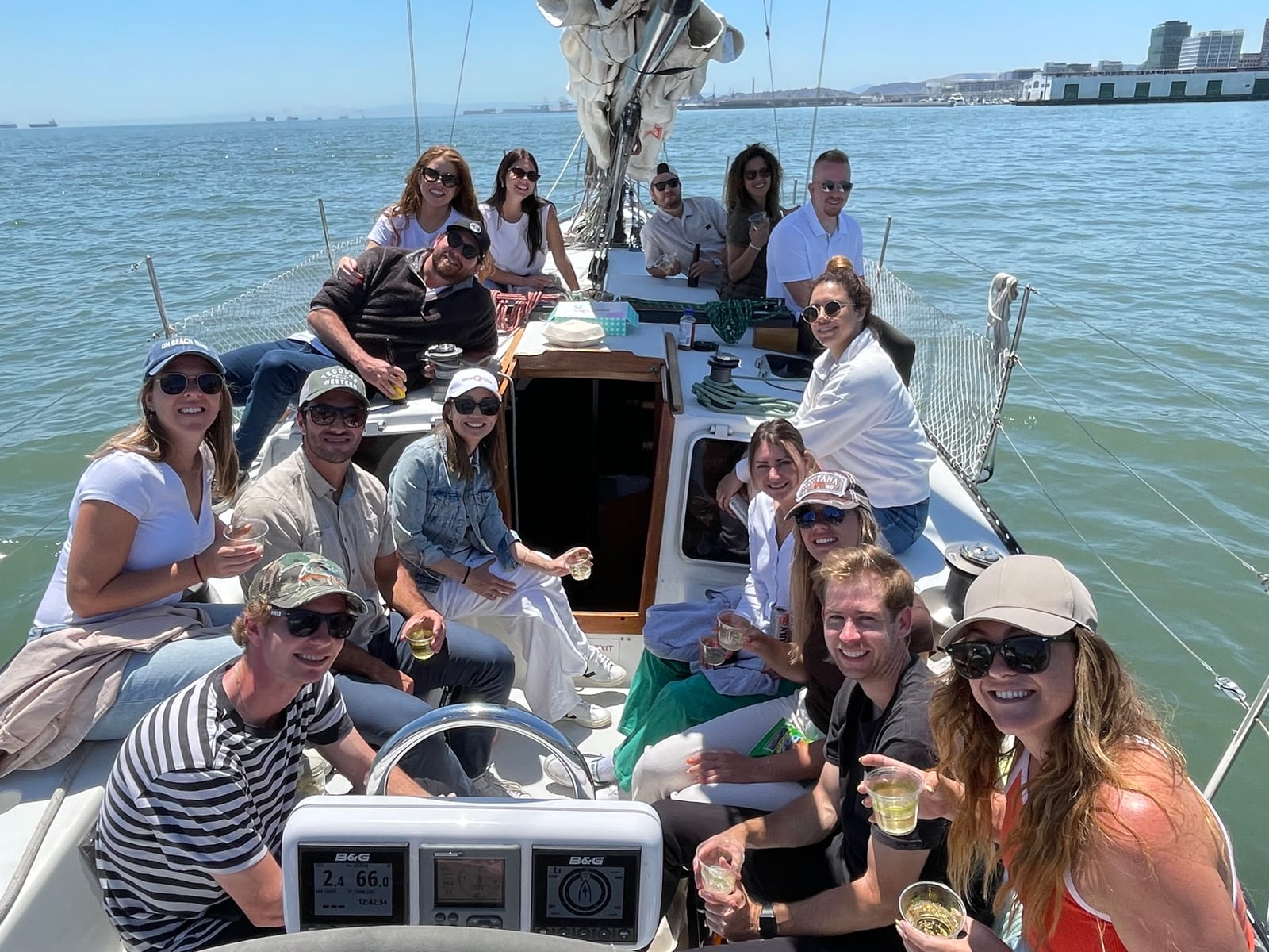 About fifteen guests sitting around the cockpit and wheel of a sailboat, smiling at the camera, with the San Francisco waterfront behind