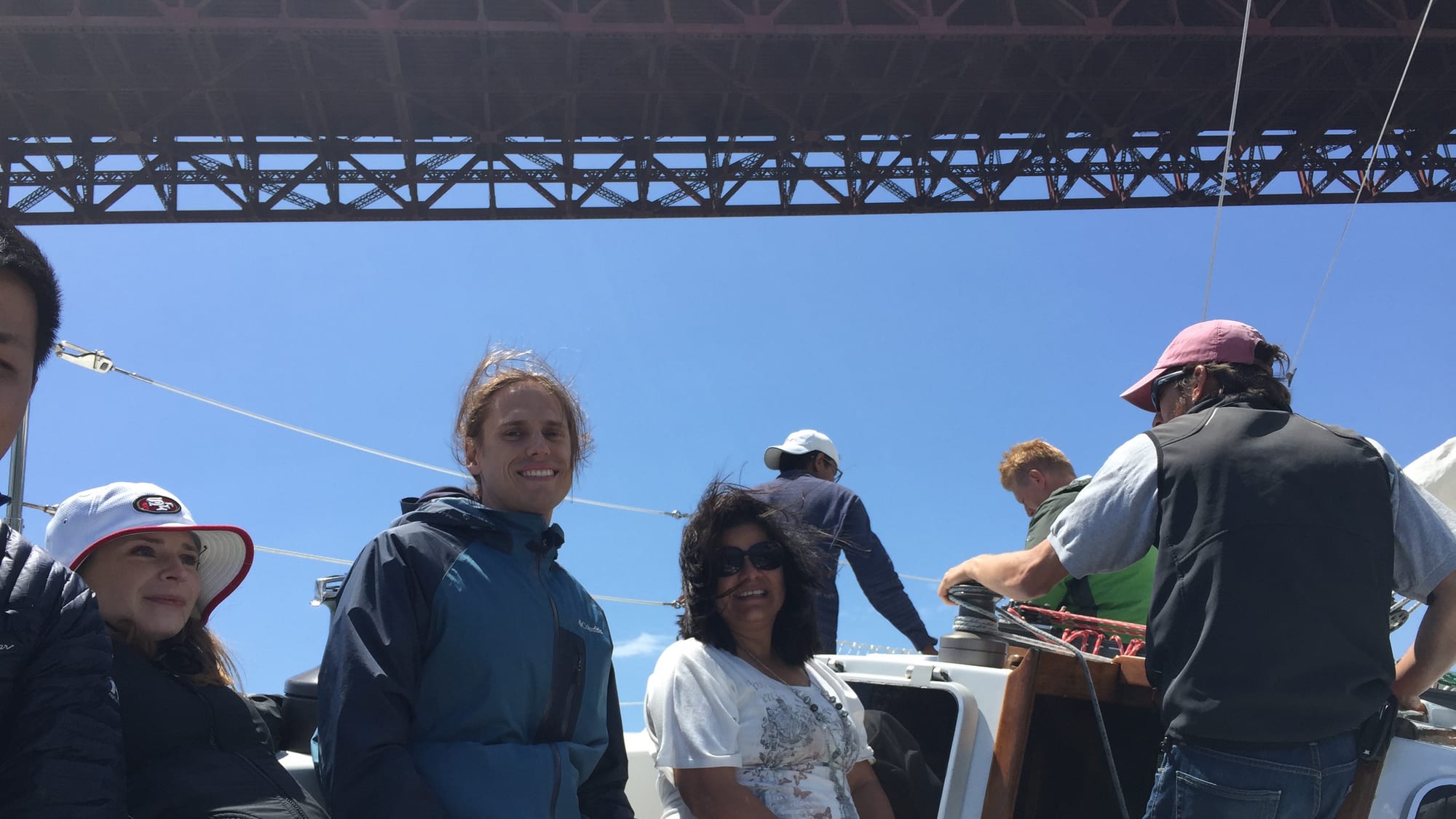Guests sitting along the cockpit of the sailboat Yukon Jack directly beneath the orange steel deck of the Golden Gate Bridge, while crew work a winch.