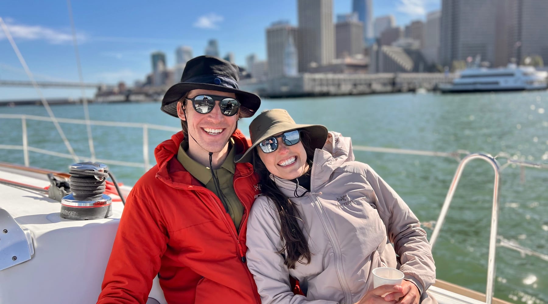 A smiling couple in sun hats, sunglasses and windproof jackets sitting on the deck of a sailboat, with the San Francisco skyline and the Ferry Building behind them