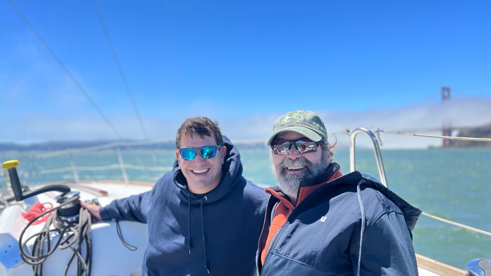Two smiling men in hoodies and jackets aboard a sailboat under blue sky, with a bank of fog swallowing a tower of the Golden Gate Bridge behind them.