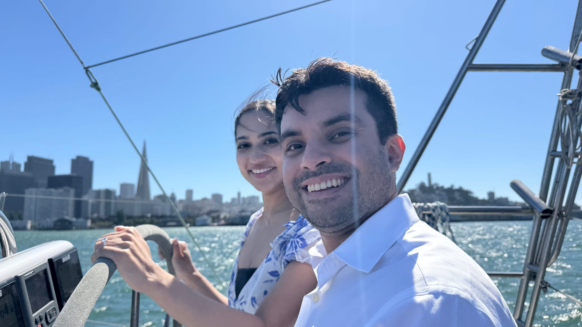 A smiling couple standing at the wheel of a sailboat on San Francisco Bay, with the Transamerica Pyramid, Coit Tower and the city skyline behind them