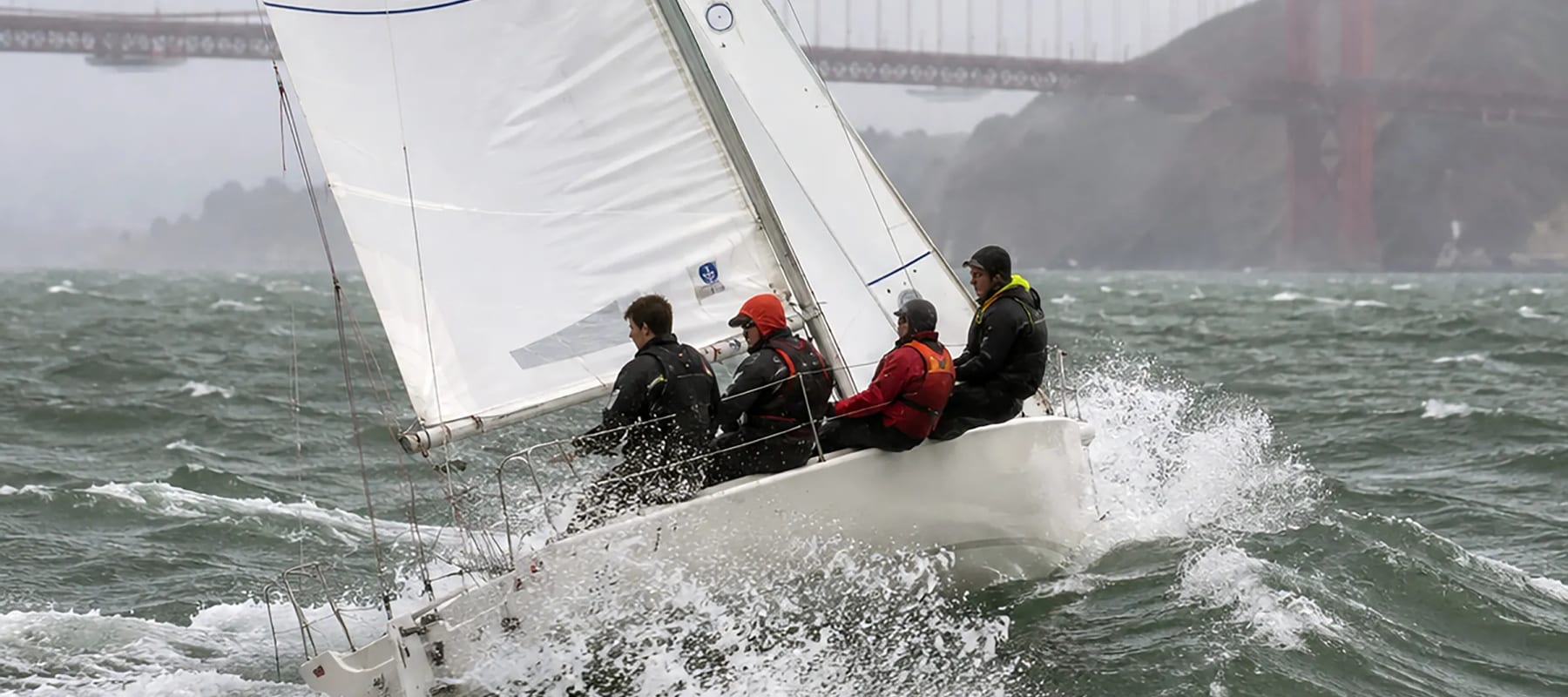 A small keelboat heeled hard over in strong wind with spray flying, the San Francisco skyline behind
