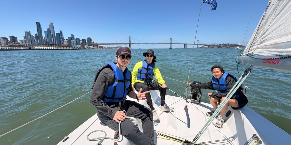 Three Spinnaker Sailing students on the bow of a J/80 with the San Francisco skyline and Bay Bridge behind them